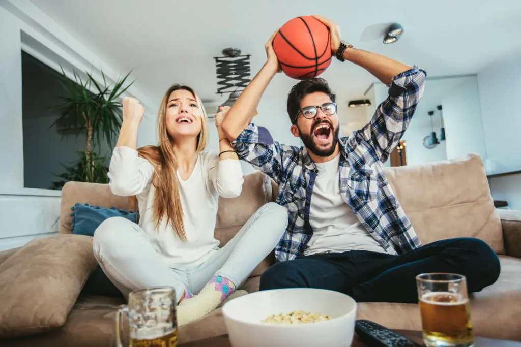 A male and female are watching basketball on a couch. March Madness fans are not only watching multiple games with Xfinity Multiview but keeping up with their March Madness brackets and scores only with Xfinity.