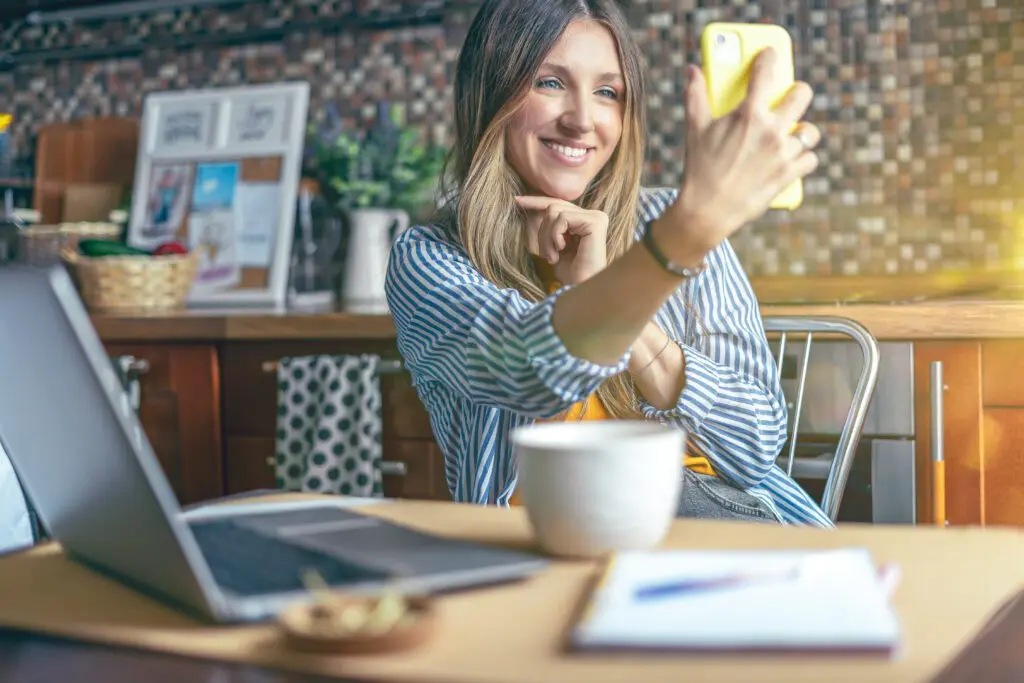 Xfinity Mobile customer in Kentucky utilizing new Premium Unlimited plans 4K streaming and hotspot data on WiFi. IMAGE: Business woman having a facetime video call. Woman make selfie. Happy and smiling girl working from home office kitchen. Using computer and mobile phone.