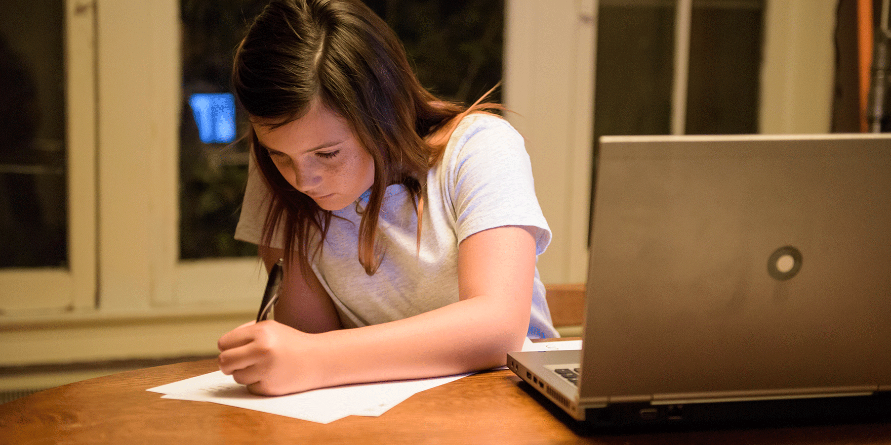 A young student writes on a piece of paper next to her laptop. Xfinity's Internet Essentials offers a low-cost internet and mobile plan.